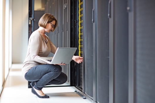 An IT professional holding a laptop, kneeling in front of a larger computer server, performing maintenance or routine check